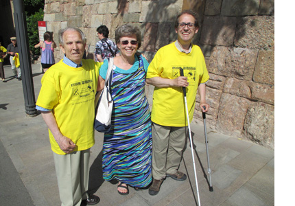 Photo shows Dona standing with two men who are wearing yellow t-shirts, Vicente is holding a white cane and a forearm crutch.